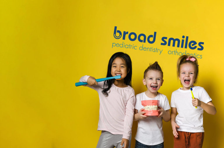 Three pediatric dental patients holding toothbrushes and a model of teeth stand in front of the Broad Smiles Pediatric Dentistry logo on a wall
