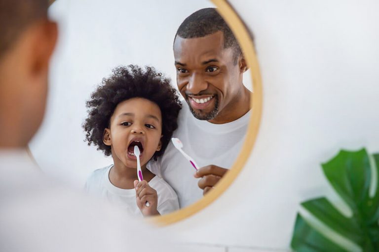 A father and child in front of a mirror practicing good oral hygiene together by brushing teeth